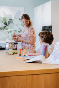Une femme souriante prépare des ingrédients dans une cuisine moderne, tandis qu'un enfant dessine avec des crayons colorés sur la table.