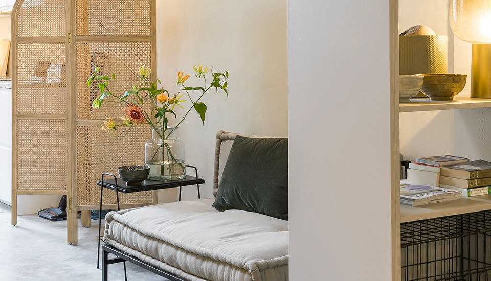 Un canapé beige avec coussin vert est placé près d'une table noire avec un vase de fleurs, séparé par un paravent en rotin.