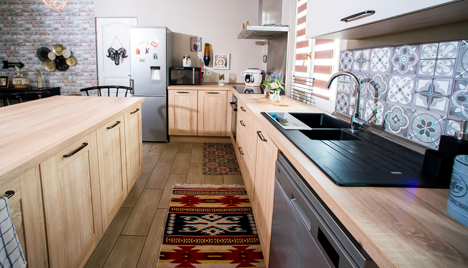 Cuisine moderne avec meubles en bois clair, plan de travail noir, carrelage à motifs et réfrigérateur argenté, éclairée par une fenêtre.