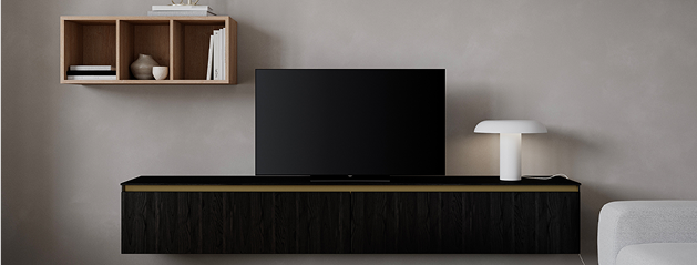 Meuble TV noir avec écran plat, lampe blanche et étagère murale en bois avec livres et vase, sur fond de mur gris.