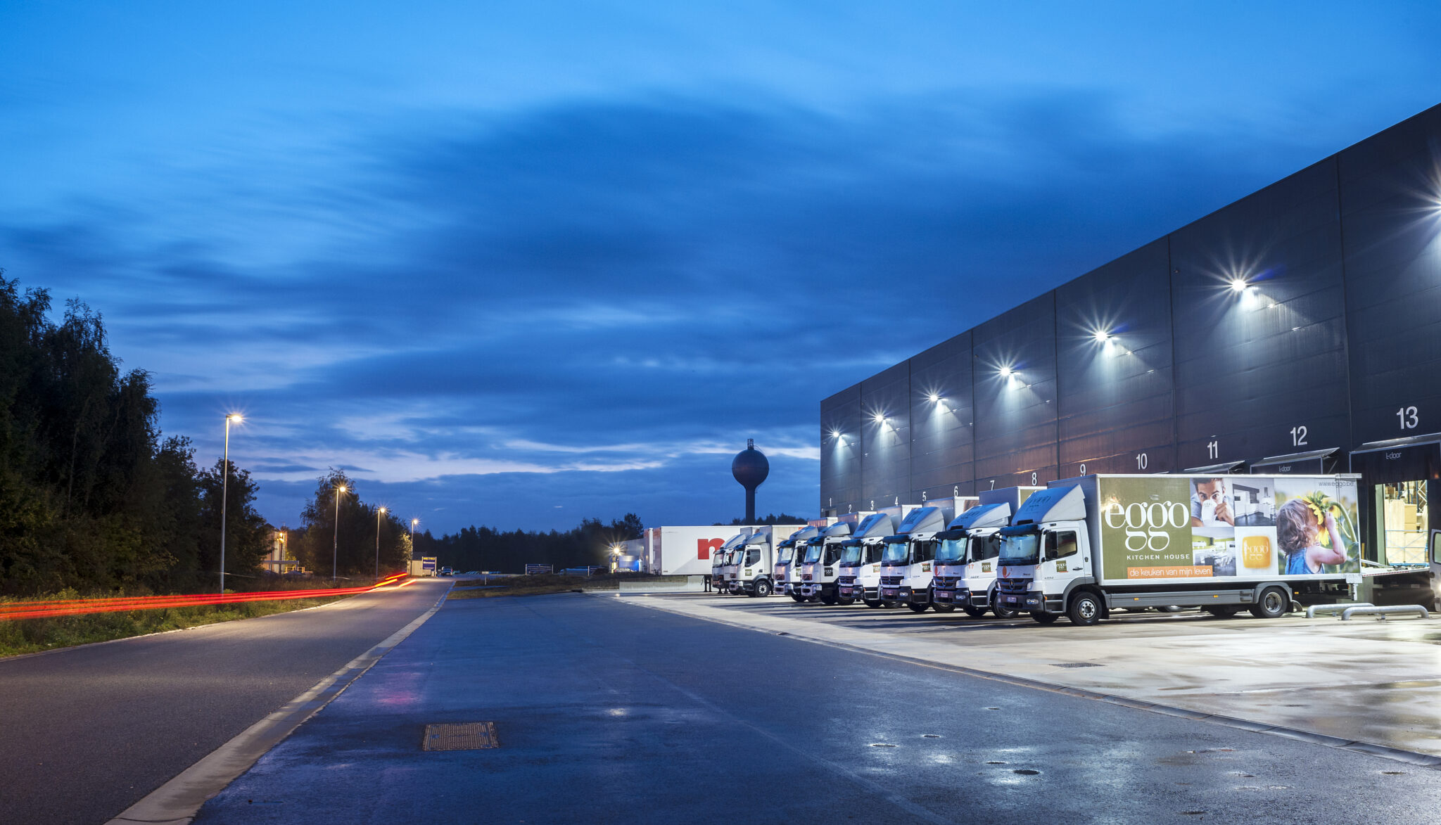 Camions alignés devant un entrepôt éclairé, sous un ciel bleu nocturne, avec des lampadaires illuminant la route à gauche.