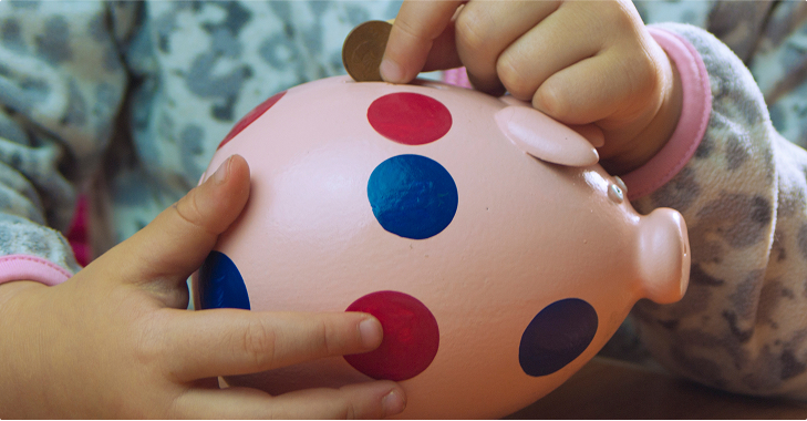 Des mains d'enfant insèrent une pièce dans une tirelire rose à pois rouges et bleus.