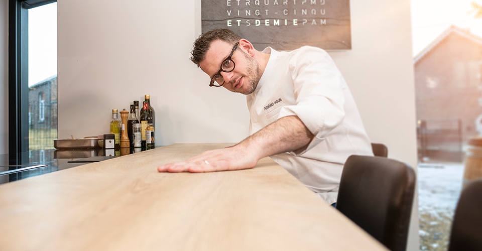 Un homme en chemise blanche touche une table en bois clair, avec des bouteilles d'huile et de vinaigre en arrière-plan.