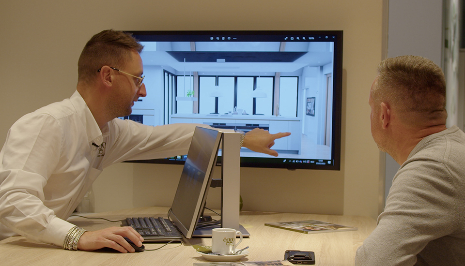 Deux hommes discutent devant un écran affichant une cuisine moderne, avec un clavier, une tasse et un magazine sur la table.