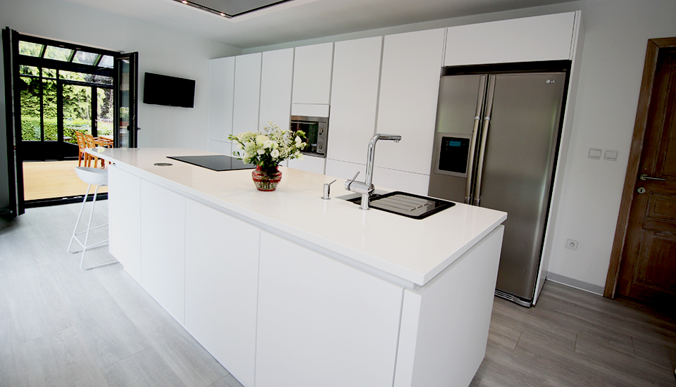 Cuisine moderne avec îlot central blanc, évier intégré, vase de fleurs blanches, et réfrigérateur en acier inoxydable à droite.