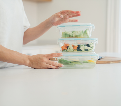Des boîtes en verre transparentes empilées contiennent des légumes coupés, posées sur un plan de travail blanc avec une main à proximité.