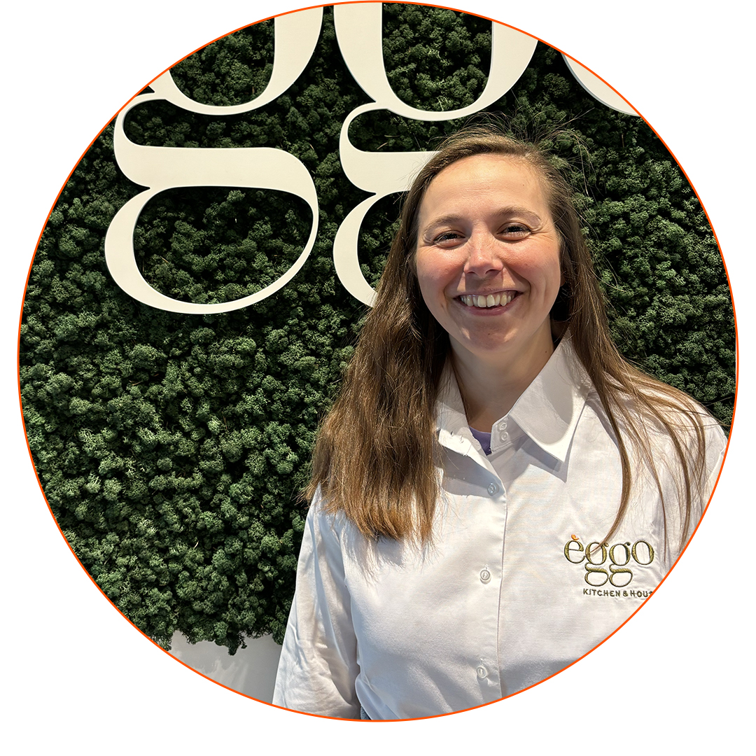 Une femme souriante en chemise blanche èggo se tient devant un mur végétal avec un logo blanc.