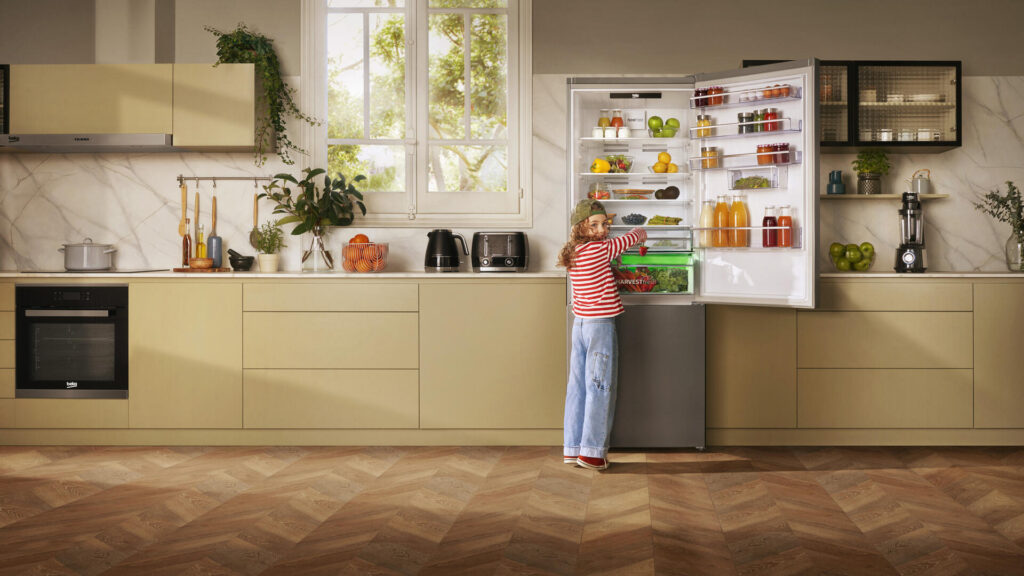 Frigo Beko ouvert et bien rempli, avec une petite fille à pull rayé, jeans et casquette qui ouvre le bac à légumes. Cuisine aux façades vertes  et mates et crédence effet marbre.