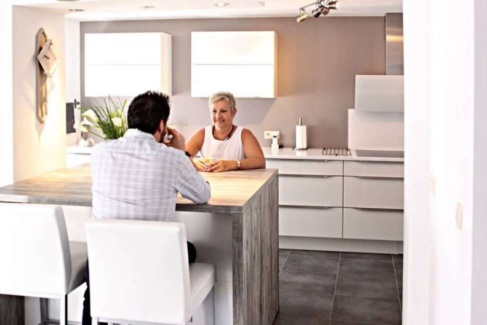 Deux personnes discutent dans une cuisine moderne avec des meubles blancs, un plan de travail en bois et des chaises blanches.