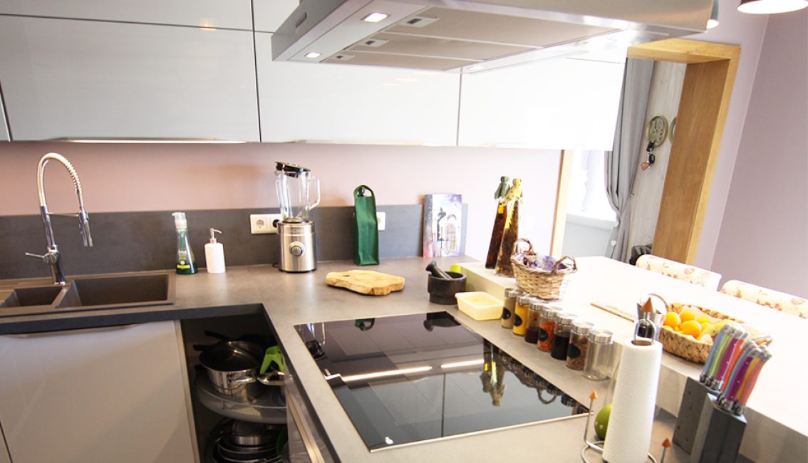 Cuisine moderne avec plan de travail gris, évier en inox, mixeur, épices alignées, planche en bois et corbeille de fruits sur la table.