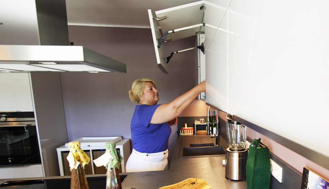 Une femme en haut bleu ouvre un placard blanc dans une cuisine moderne avec un plan de travail gris et un mixeur.
