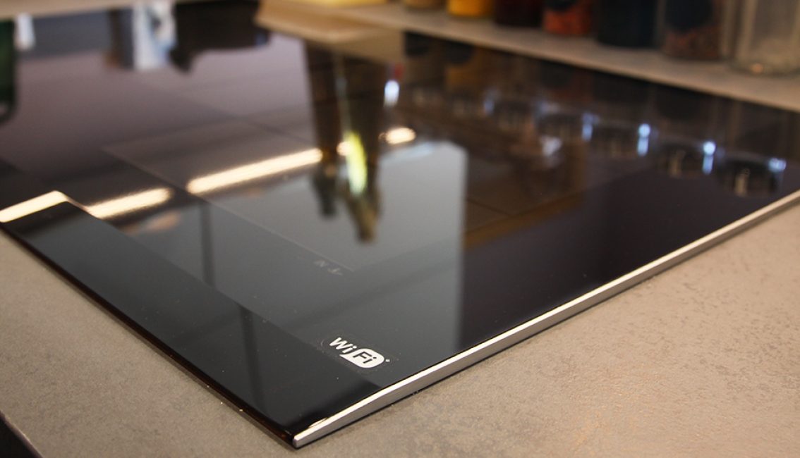 Plaque de cuisson à induction noire avec logo WiFi en coin, posée sur un plan de travail gris, avec des bocaux en arrière-plan.