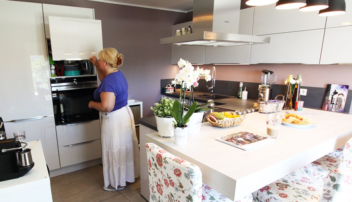 Une femme en blouse bleue et jupe blanche ouvre un placard dans une cuisine moderne avec un îlot central décoré de fleurs et de fruits.