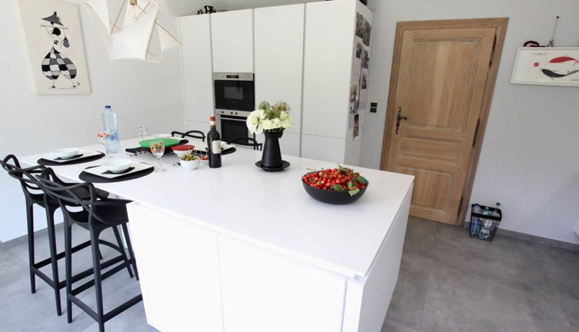 Une cuisine moderne avec un îlot central blanc, des chaises noires, un vase de fleurs, et un bol de fruits rouges.