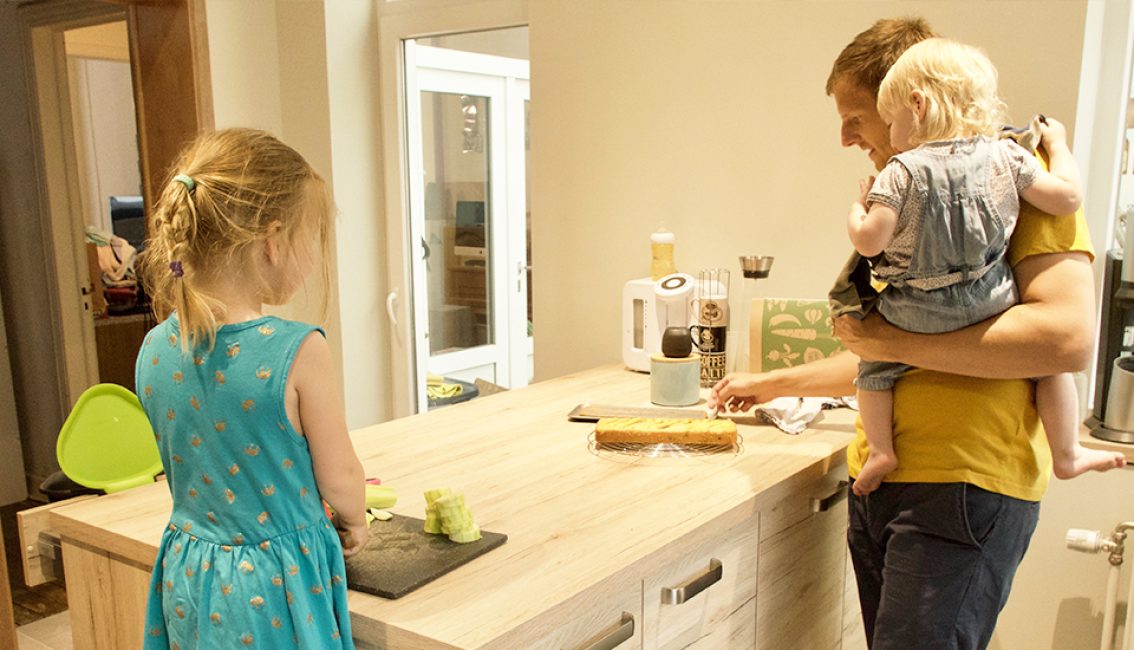 Un homme en t-shirt jaune tient un enfant blond dans une cuisine, tandis qu'une fille en robe bleue se trouve près d'un îlot central en bois clair.
